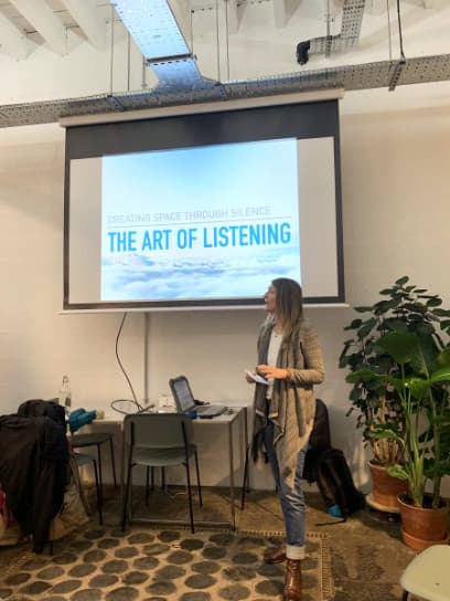 A woman standing in front of a big screen which shows a slide with The Art of Listening written on it.