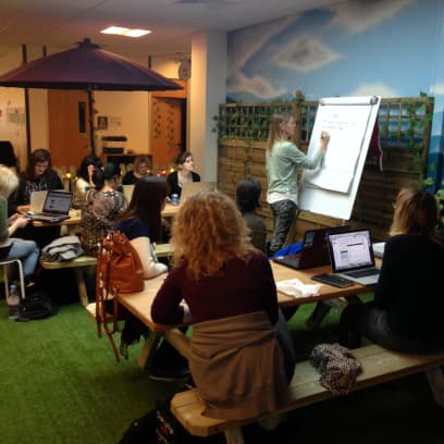 A woman standing in front of a flip chart writing. A number of women are sitting at the tables with laptops